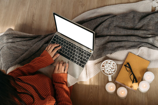 Top view of unrecognizable woman wearing a knitted sweater using laptop with copy space on the screen in home interior background. Mockup can be used to add any advertisement, texts, and graphics