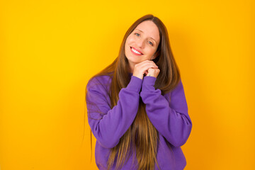 Charming serious Young beautiful Caucasian woman wearing purple sweater against yellow wall  keeps hands near face smiles tenderly at camera