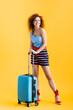 © LIGHTFIELD STUDIOS - full length of positive woman in summer outfit and wedge sandals standing near suitcase on yellow