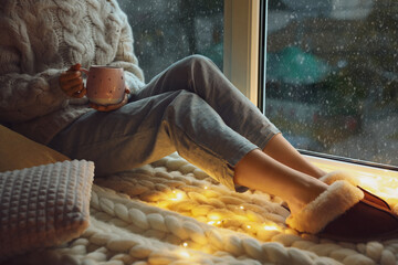 Wall Mural - Woman with drink sitting on soft plaid near window in evening, closeup