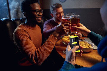 Clinking. Excited fans in bar with beer and mobile app for betting, score on their devices. Screen with match results, emotional friends cheering. Gambling, sport, finance, modern techn concept.