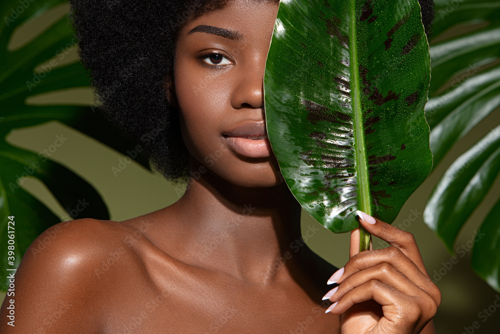 Beauty portrait of young beautiful african american woman with posing with banana leaf curly hair against green exotixc plants  background. Natural skin care concept - obrazy, fototapety, plakaty 