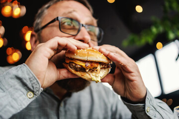 Middle aged bearded hungry man sitting in restaurant and eating delicious burger.