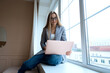 © natapetrovich - Young female in glasses focusing on screen and interacting with laptop while sitting on window sill near green potted tree in loft office. High quality photo
