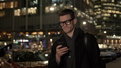 Fototapete - Man in eyeglasses with phone in hand on background of building with lights on. Handheld night shot of caucasian man texting in phone near business building with cars