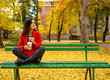 © Branimir - Woman in red coat sitting on green old wooden bench. Around her are colorful fallen leaves. She is holding cup of coffee to go.