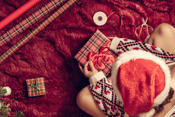 Wall Mural - Top view of woman in red santa claus hat wrapping christmas gifts boxes for family sitting on fluffy plaid near christmas tree. Flat lay.