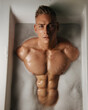 Portrait of handsome young guy with six pack abs lying in bath with foam
