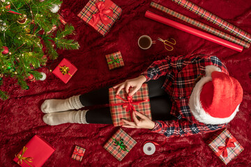 Wall Mural - Top view of woman with christmas gift box on knees. Female wrapping gifts for family sitting on fluffy plaid near christmas tree. Flat lay.