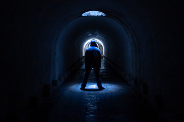 A hooded man walking towards a lighted exit from a dark, gloomy underground corridor. Light at the end of the tunnel leading to the exit to freedom.