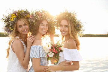 Wall Mural - Young women wearing wreaths made of beautiful flowers near river on sunny day