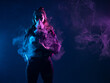 © Grispb - Vape girl releases steam from her nose. She is holding a vape device in her hand. portrait of vape girl standing in dark. Smoking electronic cigarettes. Woman vaper in cigarette smoke clubs.