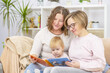 © Ermolaeva Olga - Two young women are sitting in the living room on the couch with a child on their knees and are reading a book. Gay lesbian couple at home with a baby. Lesbian family concept