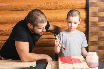 Father and son together make a wooden birdhouse in the workshop. Cheerful father with a little boy paint a wooden board with a brush in red  on a table in the workshop