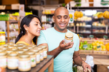 Interested Latin American Couple Reading Product Label On Jar While Choosing Groceries In Supermarket