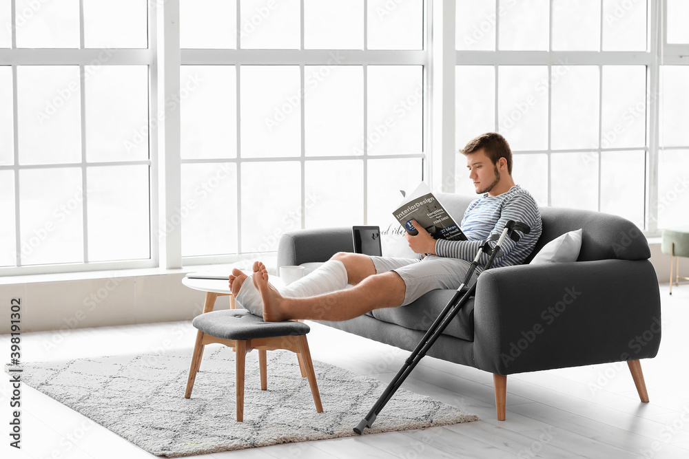 Young man with broken leg reading magazine at home