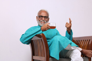 Indian old man having green tea while sitting on chair.