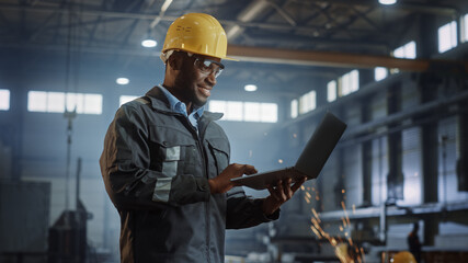 Professional Heavy Industry Engineer/Worker Wearing Safety Uniform and Hard Hat Uses Laptop Computer. Smiling African American Industrial Specialist Standing in a Metal Construction Manufacture.