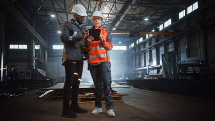 Wall Mural - Two Heavy Industry Engineers Stand in Steel Metal Manufacturing Factory, Use Digital Tablet Computer and Have a Discussion. Black African American Industrial Specialist Talk to Female Technician.