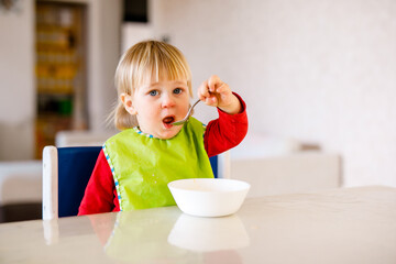 Cute baby 1,4 years old sitting on high children chair and eating vegetable alone in white kitchen