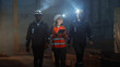 © Gorodenkoff - Three Diverse Multicultural Heavy Industry Engineers and Workers in Uniform Walk in Dark Steel Factory Using Flashlights on Their Hard Hats. Female Industrial Contractor is Using a Tablet Computer.