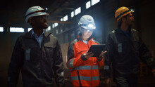 Three Diverse Multicultural Heavy Industry Engineers And Workers In Uniform Walk In Dark Steel Factory Using Flashlights On Their Hard Hats. Female Industrial Contractor Is Using A Tablet Computer.