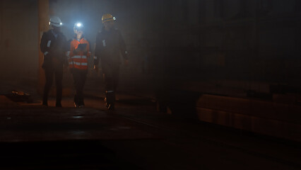 Wall Mural - Three Diverse Multicultural Heavy Industry Engineers and Workers in Uniform Walk in Dark Steel Factory Using Flashlights on Their Hard Hats. Female Industrial Contractor is Using a Tablet Computer.