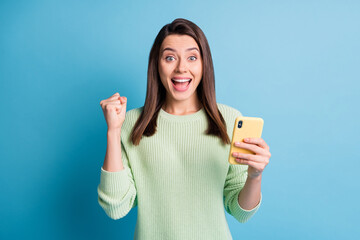 Wall Mural - Photo portrait of excited girl celebrating with fist in air holding phone in one hand isolated on pastel blue colored background