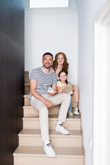 Wall Mural - Happy family with teddy bear and statuette of house looking at camera while sitting on stairs at home