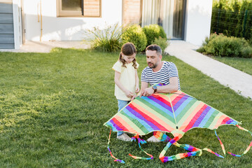 Wall Mural - Full length of daughter standing near father assembling kite on lawn with house on background