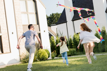 Wall Mural - Back view of family running while flying kite on lawn near home