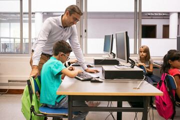 Positive male school teacher helping boy to cope with his task in computer science class. Side view. Education or back to school concept