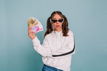 Fashionable girl with collected hair wearing black sunglasses and white pullover posing with a wad of money on isolated background