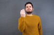 © Roquillo - Young handsome Caucasian man wearing yellow sweater against gray wall angry gesturing typical italian gesture with hand, looking to camera