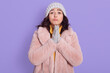 © sementsova321 - Attractive cute European girl in fur coat, cap and mittens, prays on lilac background, keeping palms together, asking for something important, poses with pout lips.