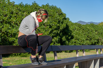 Sticker - Shot of a  woman in a protective mask with headphones  standing on a bench in a park