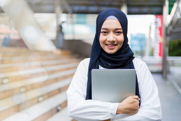 Islamic student or working woman standing with her arms crossed holding her laptop, smiling confidently.