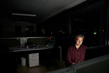 Middle aged businessman is sitting at a laptop in a dark office working on new deadline project - young male working late at night in the office