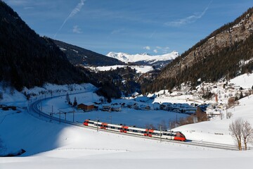 Wall Mural - A train traveling through the railway curve in a valley covered by heavy snow and St. Jodok village in Brenner Pass lying on the hillside of Alpine mountains on a sunny winter day, in Tyrol, Austria