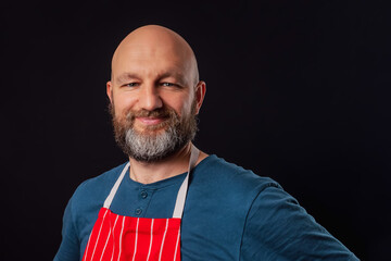 Portrait of professional butcher or fishmonger in blue t shirt and red and white stripe classic apron on a black background. The model is bald with grey and black beard in his 40s. Smiling expression