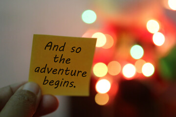 New year inspirational motivational quote on a paper note in hand - And so the adventure begins. On blurry background of colorful Christmas light bokeh. Happy New Year.