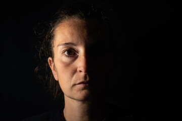 Dark portrait of a serious woman with only half face illuminated on black background. Feelings of loneliness, depression and anxiety.