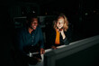 © StratfordProductions - Two smiling young multiethnic business colleagues sitting together at desk working late at night in dark office on computer - two colleagues finishing up deadlines for months end