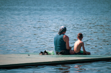 Father and son fishing on the lake. Father and son are sitting on a small fishing pier, holding sticks, their backs facing the camera. Family vacation on the water. A father teaches his son to fish.