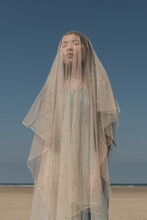 Girl On Beach Covered By Thins White Fabric Like A Virgin Mary Statue Under A Blue Sky
