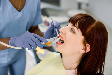 Wall Mural - Close-up side view face portrait of pretty young Caucasian woman patient having dental treatment in dentist office. Hands of african dentist in gloves holding dental tools