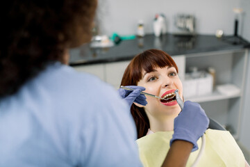 Wall Mural - Close up of young beautiful lady with healthy white teeth, sitting in dental chair, while black female dentist cures her teeth. Back view of dentist wearing latex gloves and holding tools in hands