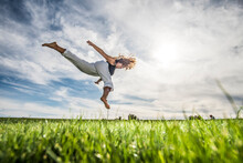 Female Athlete Jumping In Mid-air Over Grass At Park Against Cloudy Sky