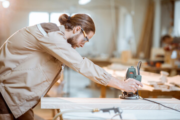 Wall Mural - Handsome carpenter in uniform chamfers wooden bar with a hand machine at the carpentry manufacturing