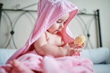 Baby Girl In Pink Towel Playing Bath Sponge While Sitting On Bed In Bedroom